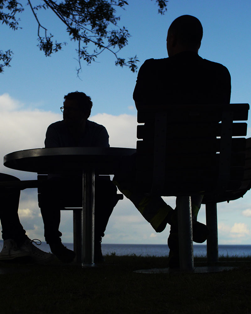 People sit on outdoor benches against the setting sky, they are silhoutted.